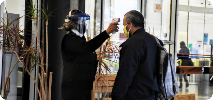 Um empregado da empresa, vestindo roupa preta, está em pé na entrada da sede. Ele usa máscara transparente cobrindo todo o rosto e aponta um termômetro digital para a testa de um visitante, que veste roupas escuras e usa máscara branca. 